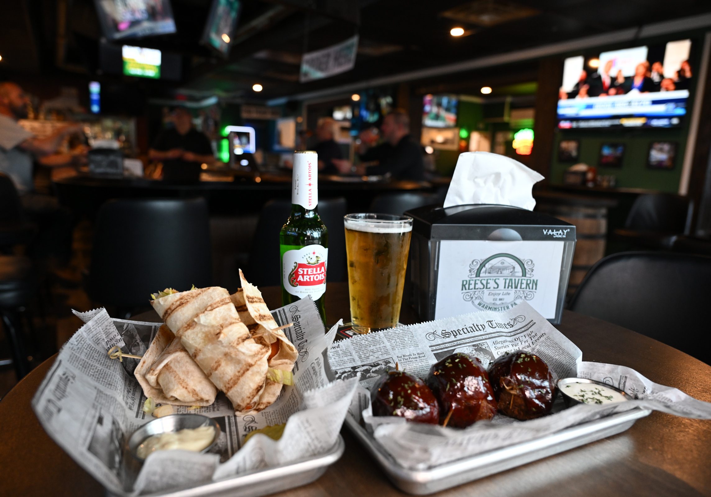 A table at a sports bar with pita bread, sauce, glazed meatballs, a bottle of Stella Artois, a glass of beer, and a Reese's Tavern napkin holder; people and TVs in the background.