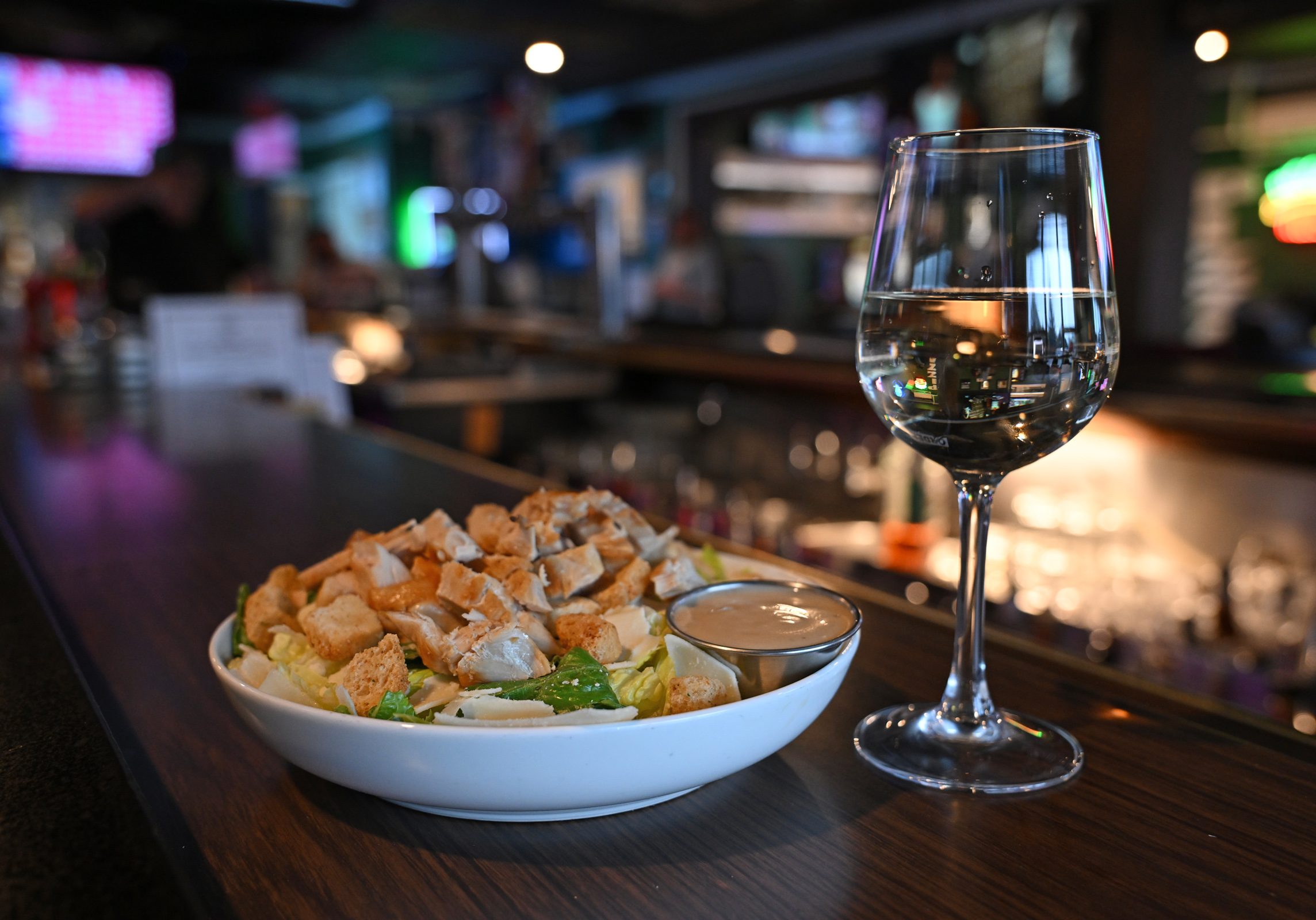 A bowl of Caesar salad with dressing and a glass of white wine are set on a bar counter in a dimly lit restaurant.
